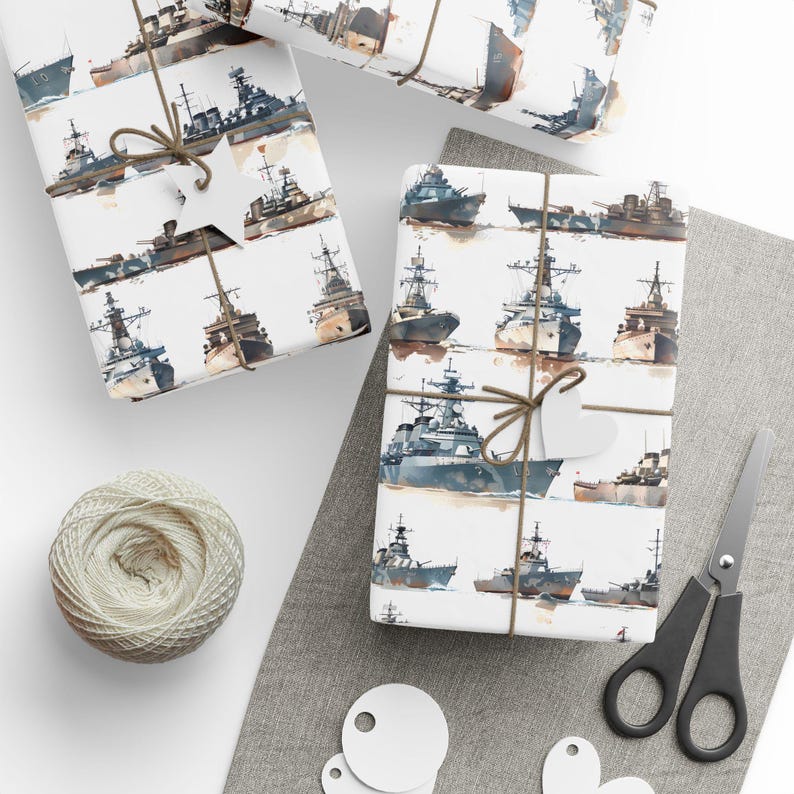 A wrapped gift box featuring watercolor warships patterned on white paper, placed on a gray mat next to a ball of twine, scissors, and blank tags.