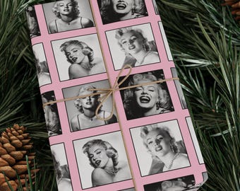May include: Pink wrapping paper featuring black and white images of Marilyn Monroe in a grid pattern. The paper is tied with twine. The background includes pine needles and a pine cone.