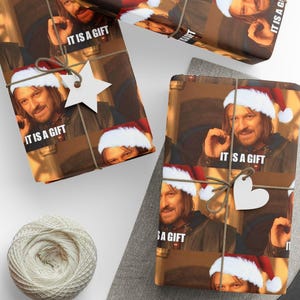 May include: Wrapping paper featuring a repeating image of a man with long brown hair wearing a red and white Santa hat, with the text IT IS A GIFT printed below. The paper is tied with brown twine and includes a white star or heart-shaped gift tag.