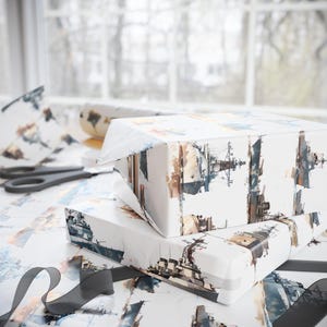 Two wrapped gift boxes featuring a battleship watercolor pattern resting on a surface with a blurred window background, scissors, and a black ribbon.