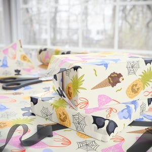 May include: Two wrapped gifts with cream-colored paper featuring Halloween-themed illustrations. The design includes pineapples with jack-o'-lantern faces, ice cream cones, spiderwebs, bats, and sunglasses. A pair of scissors and a ribbon are also visible.