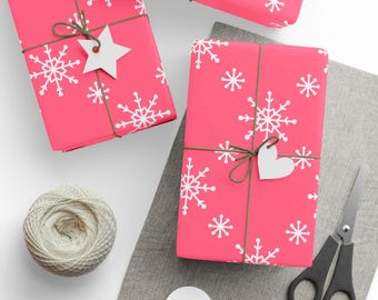 May include: Overhead view of wrapped presents in vibrant pink paper, decorated with white snowflake designs. The gifts are tied with brown twine and have white paper tags shaped like stars and hearts. A ball of twine, scissors, and extra tags are also present.