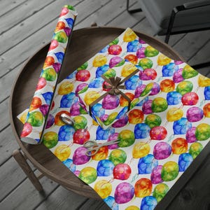 A round wooden table displays an open sheet of watercolor rainbow balloon wrapping paper, a matching wrapped gift box, and a roll of the same paper.