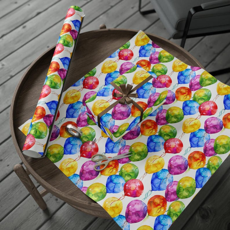 A round wooden table displays an open sheet of watercolor rainbow balloon wrapping paper, a matching wrapped gift box, and a roll of the same paper.