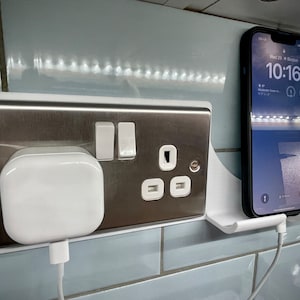 May include: A white phone charging stand with a white phone charger plugged into a white electrical socket. A white smartphone is mounted on the stand and the screen shows the time 10:16. The phone is charging.