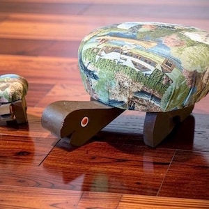 May include: A large and a small turtle-shaped footstool, each featuring a dark brown wooden base and a padded seat upholstered in fabric with a multi-colored landscape print. The turtle heads are brown with a single white and orange circular eye detail.