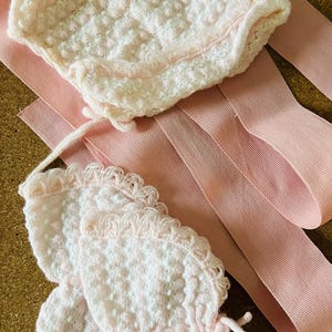 May include: A vintage-style baby bonnet and matching booties set in soft pink. The bonnet and booties are made of a textured, possibly crocheted, material with delicate trim. A pink ribbon is also included.