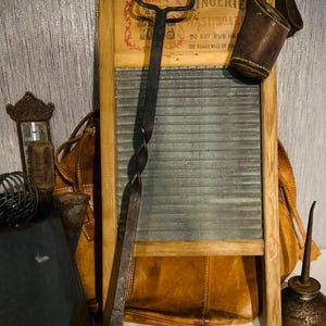 May include: An assortment of vintage items, including a wooden washboard with a metal scrubbing surface, a leather pouch, a twisted metal tool, and an oil can. The washboard has text that reads "INGENIE WASHBOARD". The items are arranged on a white surface.