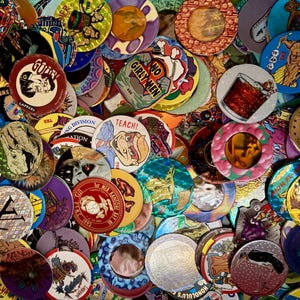 May include: A large collection of circular cardboard pogs and metal slammers featuring various colourful graphic illustrations, cartoon characters, text, and holographic patterns, layered together in a dense, overlapping pile.