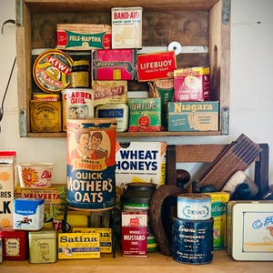 Pode incluir: Uma coleção de recipientes vintage de alimentos e produtos domésticos, incluindo caixas de Band-Aid, sabonete Lifebuoy e Quick Mother's Oats. Os itens são exibidos em uma prateleira e mesa de madeira, evocando uma sensação de nostalgia.