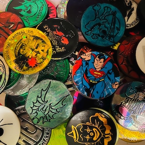 May include: A collection of circular and octagonal pogs featuring various graphic designs, including a Superman illustration, a clown, a spider, a skull, and a Super Bowl XXX logo. The pogs display diverse colors, holographic finishes, and text.