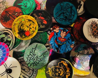 May include: A collection of circular and octagonal pogs featuring various graphic designs, including a Superman illustration, a clown, a spider, a skull, and a Super Bowl XXX logo. The pogs display diverse colors, holographic finishes, and text.