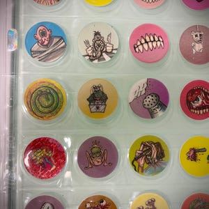 May include: A collection of round, colorful collectible pogs with various cartoon monster and creature designs. The pogs are displayed in a clear plastic sheet, showcasing the diverse artwork and vibrant colors of each pog.