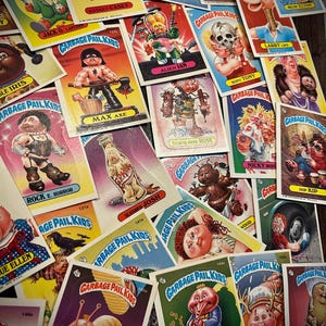 May include: A collection of vintage Garbage Pail Kids trading cards. The cards feature colorful, cartoonish characters with humorous and often grotesque designs. The cards are scattered across a dark surface, showcasing various characters and card titles.