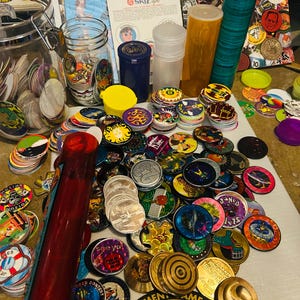 Peut inclure: Une collection de pogs colorés, de capsules de bouteilles et de pièces de jeu. L'image comprend une variété de motifs, notamment des personnages de dessins animés, des logos et des motifs abstraits. Il y a aussi des contenants, un bocal et un objet rouge en forme de batte.