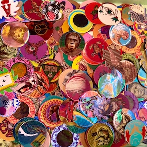Peut inclure: Une grande collection de pogs circulaires en carton présentant divers motifs graphiques, notamment des illustrations d'animaux, de visages humains, de motifs abstraits et de cartes à jouer dans des couleurs vives et des finitions holographiques.