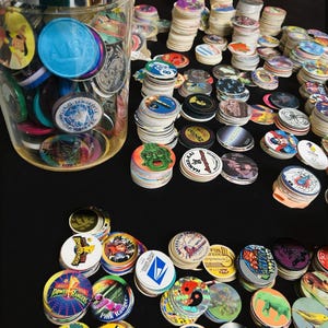 May include: A collection of colorful pogs, some stacked and others scattered, with various designs and text. A glass jar filled with pogs is on the left. The pogs feature images of Power Rangers, cartoon characters, and other graphics.