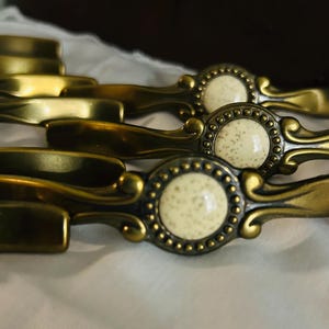 May include: Close-up of several antique brass drawer pulls. Each pull features a decorative design with a central cream-colored, speckled insert surrounded by a beaded border. The pulls have a curved handle and are arranged in a row.