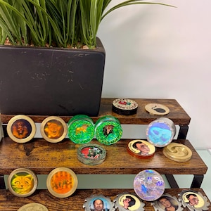 May include: A collection of circular decorative coins and resin plugs featuring various designs including portraits, animals, and holographic patterns. The items are arranged on a tiered wooden display stand with metallic and glitter finishes.