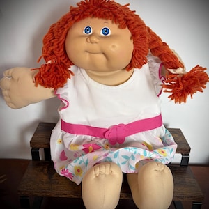 May include: A vintage doll with red yarn hair styled in braids. The doll has blue eyes and is wearing a white dress with a pink belt and floral skirt. The doll is sitting on a dark brown wooden bench.