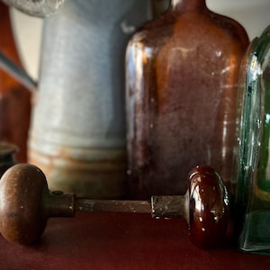 Puede incluir: Un pomo de puerta de madera vintage con un mango de cerámica marrón oscuro está en primer plano. Detrás hay varias botellas antiguas y una regadera de metal. Las botellas son de varios colores, incluyendo marrón y verde.