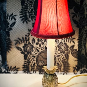 May include: A small, decorative lamp with a red lampshade and a gold pineapple base. The lampshade is a deep red with a velvet trim. The base is a detailed gold pineapple design. The lamp is lit, casting a warm glow.