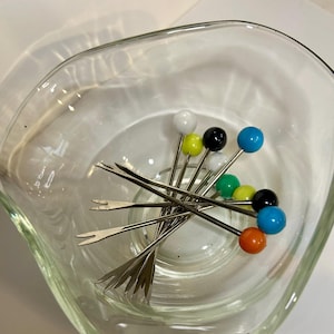 May include: A clear glass serving bowl holding a collection of cocktail picks. The picks have silver stems and colorful ball-shaped tops in white, yellow, black, blue, green, and orange. The bowl has a unique, wavy shape.