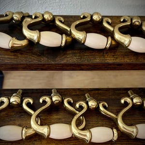 May include: Eight brass drop pulls with white oval ceramic inserts. Each pull features a curved metal handle attached to a central mounting point, showcasing a polished gold-tone finish and a smooth, opaque white center accent piece.