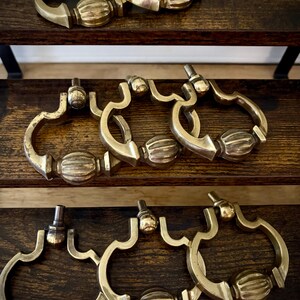 May include: A collection of eight brass drop pulls featuring a curved, ornate frame and a central ribbed, oval-shaped detail. Each pull includes a small, rounded mounting post at the top, designed for attachment to furniture or cabinetry hardware.