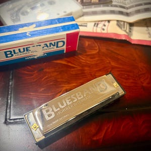 以下が含まれることがあります： 「Bluesband Hohner International」と「Made in China」の文字が入った、銀色のホーナーブルースバンドハーモニカ。ハーモニカは濃い木製の表面に置かれており、背景には箱やその他のハーモニカアクセサリーがあります。