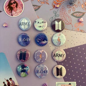 Peut inclure: Une collection de badges ronds et colorés sur le thème de la K-Pop. Les badges présentent divers motifs, notamment le mot "ARMY", l'expression "Purple you", du texte coréen et un geste de la main en forme de cœur. Le fond comprend un autocollant qui dit "BLOOM STUDIO KPOP X LUV".