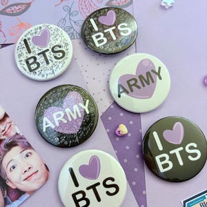 May include: A collection of round BTS-themed buttons in various colors. The buttons feature the phrases "I Love BTS" and "ARMY" with a purple heart design. Some buttons have a black background, while others are white.