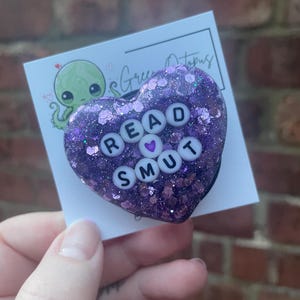 May include: Purple heart-shaped resin keychain with glitter and white letters spelling "READ SMUT" with a small pink heart between the words.