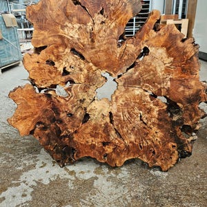 May include: A large, irregularly shaped wooden slab with a natural, organic form. The slab features a rich, warm brown color with intricate grain patterns and knots. The wood has a rough, textured surface with several holes and cracks, adding to its rustic appeal.