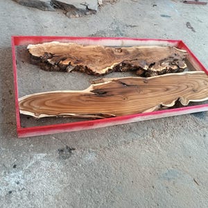 May include: Two pieces of wood are placed inside a red frame. The wood is a light brown color with dark brown grain patterns. The wood is being prepared for a resin project.
