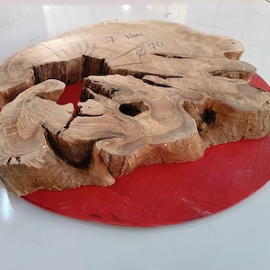 Puede incluir: Una gran rebanada de madera natural con una forma única e irregular y un agujero en el centro. La madera es de color marrón claro con marcas marrones más oscuras. La rebanada está sentada sobre una base circular roja.