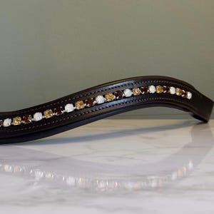 Peut inclure: Un frontal de bride de cheval incurvé en cuir marron foncé. Le frontal est orné d'une rangée de cristaux blancs, ambrés et rouge foncé alternés. Les cristaux sont disposés en ligne le long du haut du frontal.