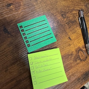 Könnte beinhalten: Eine grüne quadratische Schablone mit sechs horizontalen Schlitzen und sechs quadratischen Kästchen auf der linken Seite, über einem lindgrünen Haftnotizzettel mit handschriftlichen Aufgaben wie Send emails, Check website, Call school und Order dinner.