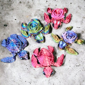 Puede incluir: Cinco pequeñas figuritas de tortugas con caparazones en forma de rosa. Las tortugas están hechas de un material brillante e iridiscente y vienen en una variedad de colores, incluyendo rosa, azul, verde y morado.