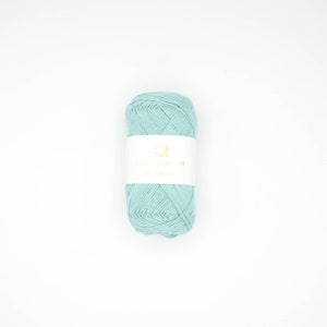 Könnte beinhalten: Ein Knäuel helltürkisfarbenes Garn mit einem weißen Etikett. Das Etikett hat goldenen Text und ein geometrisches Design. Das Garn ist fest gewickelt und wirkt weich, geeignet für Strick- oder Häkelprojekte.