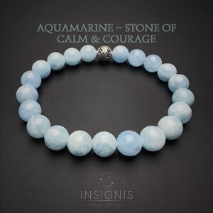 Puede incluir: Una pulsera de cuentas redondas de aguamarina azul pálido dispuestas en círculo, con una cuenta de metal plateado ornamentada en la parte superior. El texto AQUAMARINE - STONE OF CALM & COURAGE y la marca INSIGNIS son visibles.