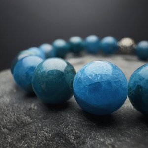 Puede incluir: Un primer plano de un brazalete de piedras preciosas azules. El brazalete está hecho de cuentas redondas y lisas de un color azul intenso. Las cuentas están enhebradas en un cordón elástico transparente.