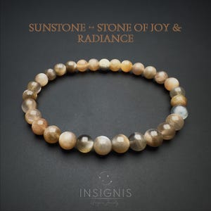 Può includere: Un braccialetto circolare di perline con pietre del sole lisce e rotonde in tonalità marrone chiaro, beige e grigio traslucido. Una singola perlina testurizzata dai toni metallici è posizionata in alto. Il testo sopra recita: SUNSTONE - STONE OF JOY & RADIANCE.