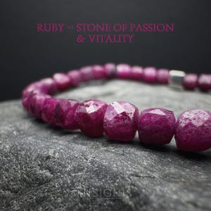 Könnte beinhalten: Nahaufnahme eines Rubinarmbands mit facettierten Perlen. Das Armband hat eine tiefmagentafarbene Farbe und liegt auf einer grauen, strukturierten Oberfläche. Der Text "RUBY - STONE OF PASSION & VITALITY" ist rosa dargestellt.