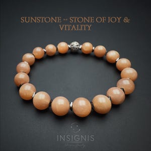 Può includere: Un bracciale composto da perle rotonde di pietra del sole color pesca, alternate a piccoli distanziatori metallici scuri e a una singola perla sferica argentata decorata. Il testo SUNSTONE - STONE OF JOY & VITALITY e INSIGNIS è visibile.