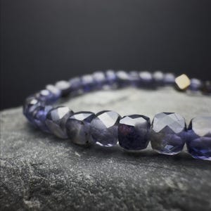 May include: A close-up of a bracelet featuring faceted, dark purple beads. The beads are strung together, with a small gold-colored square bead at the end. The bracelet is resting on a gray, textured surface.