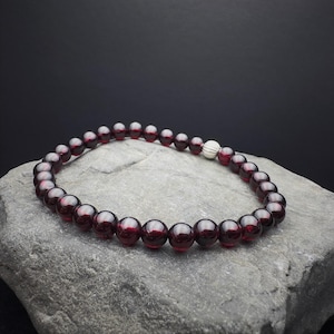 Könnte beinhalten: Ein Armband mit tiefroten, runden Edelsteinperlen. Das Armband hat eine einzelne, silberfarbene Perle. Es wird auf einem grauen, strukturierten Felsen vor dunklem Hintergrund präsentiert.