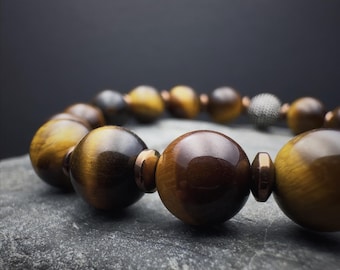 May include: A close-up of a bracelet featuring large, polished tiger's eye beads. The beads are a mix of golden brown and dark brown, with a metallic accent bead. The bracelet is resting on a textured gray surface.