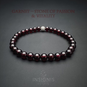 May include: A dark red garnet bead bracelet with a silver bead. The bracelet is made of polished garnet stones. Text at the top reads "GARNET - STONE OF PASSION & VITALITY". The bracelet is displayed on a dark grey background.