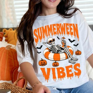 May include: A white t-shirt featuring a graphic of a skeleton wearing sunglasses, relaxing in an orange and white striped pool float. The design includes the text "SUMMERWEEN VIBES" in orange, along with illustrations of bats and jack-o'-lanterns.